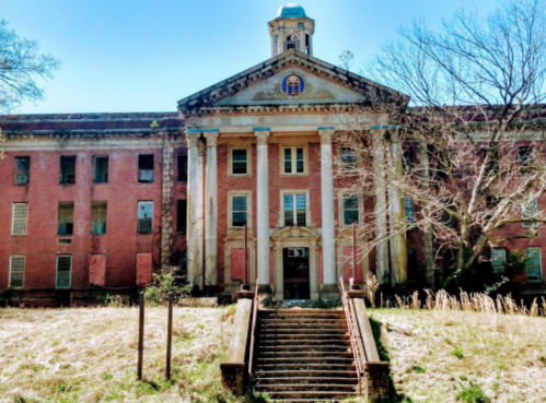 Abandoned building with a grand entrance, tall columns, and overgrown grass in front, under a clear blue sky.