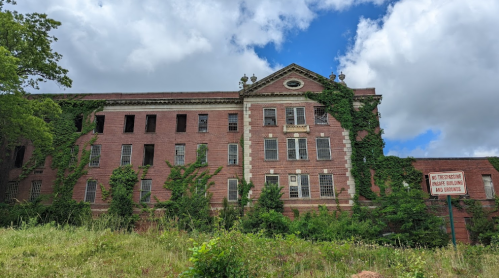 Abandoned brick building overgrown with vines, under a cloudy sky, with a "No Trespassing" sign in front.