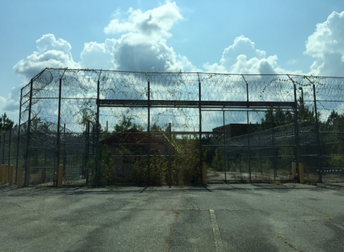 A chain-link fence topped with barbed wire surrounds an abandoned building, under a cloudy blue sky.