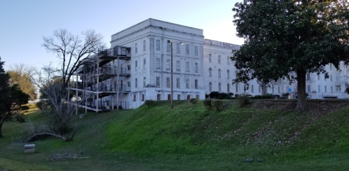 A large, historic building with a white facade, surrounded by greenery and trees, featuring a fire escape on the side.