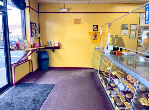 Interior of a bright yellow bakery with a display case of donuts and a counter, featuring a trash can and a welcome mat.