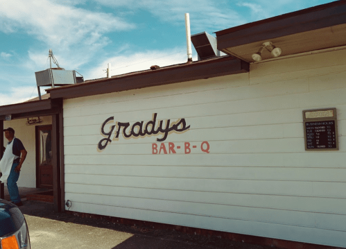 Sign for Grady's Bar-B-Q on a white building, with a person standing nearby. Clear blue sky in the background.