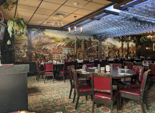 Cozy restaurant interior with red chairs, round tables, and a mural of a scenic village on the wall.