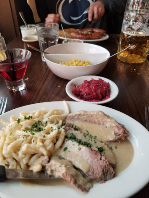 A plate of sliced meat with creamy sauce, spaetzle, and red cabbage, with drinks and other dishes in the background.