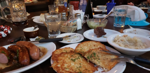 A table spread with various dishes, including sausages, hash browns, and sides, alongside drinks and utensils.