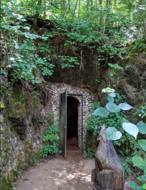 A stone doorway set into a rocky hillside, surrounded by greenery and a wooden sculpture nearby.