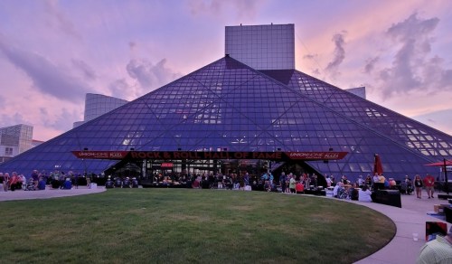 The Rock & Roll Hall of Fame, a glass pyramid structure, with a crowd gathered outside during sunset.