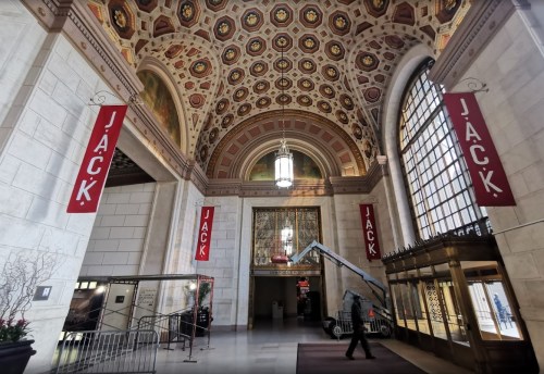 Interior of a grand hall with ornate ceiling, large windows, and banners reading "JACK." A person operates a lift.