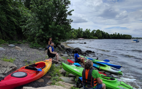 A group of people sitting by the water with colorful kayaks on the shore, surrounded by trees and a cloudy sky.