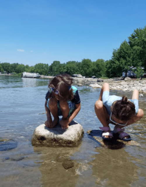 Two children crouch on rocks by a calm lake, exploring the water on a sunny day. Green trees line the shore.