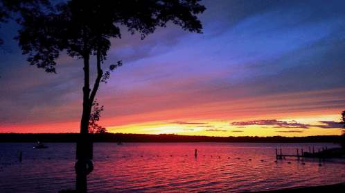 A serene lake at sunset, with vibrant colors in the sky and silhouettes of trees and boats on the water.