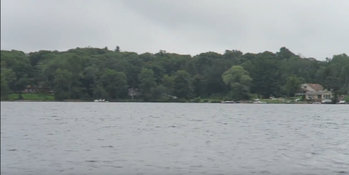 A calm lake surrounded by lush green trees under a cloudy sky, with a few boats visible in the distance.