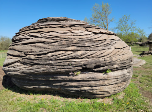 A large, rounded rock formation with layered textures, surrounded by green grass and trees under a clear blue sky.