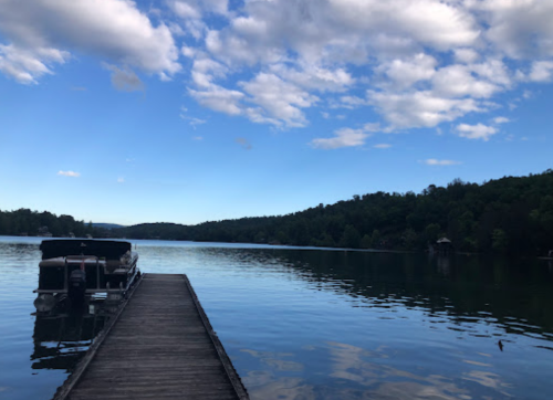 A serene lake view with a wooden dock extending into calm waters, surrounded by lush green hills and a blue sky.