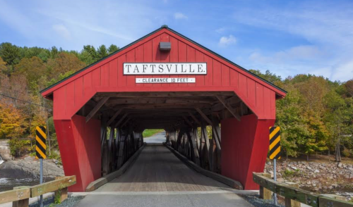 A red covered bridge with "Taftsville" sign, surrounded by trees and a clear blue sky.