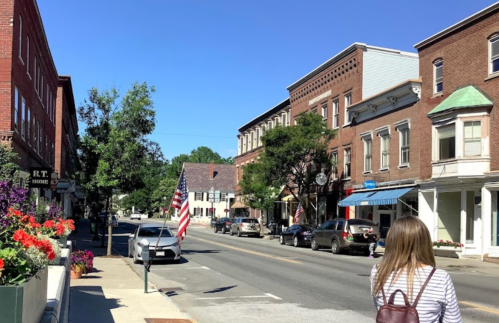 A sunny street scene with historic buildings, an American flag, and a person walking by colorful flower planters.