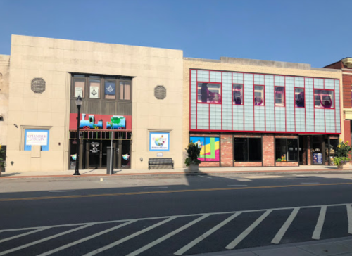 Two buildings side by side on a street, featuring colorful murals and large windows, under a clear blue sky.
