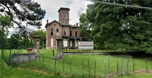 A large, old brick mansion with a tower, surrounded by trees and a fence, featuring a sign that reads "Haunt Castle."