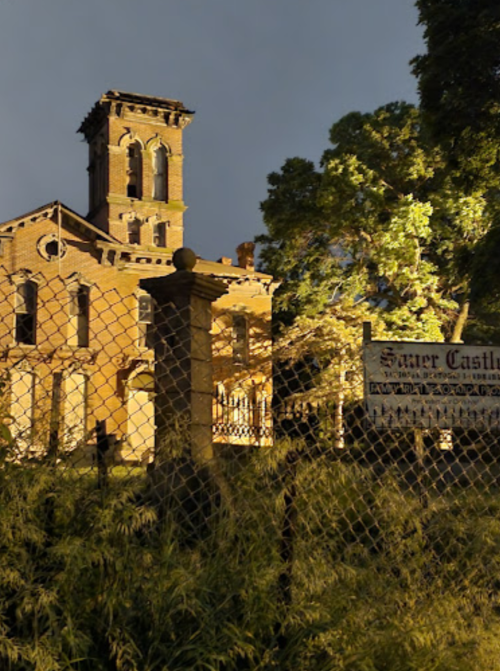 A historic, abandoned building illuminated at night, surrounded by a fence and overgrown vegetation.