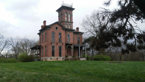 A tall, abandoned brick house with a tower, surrounded by overgrown grass and trees under a cloudy sky.