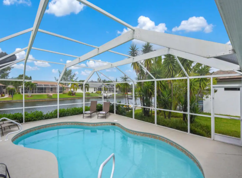 A serene pool area with a screened enclosure, surrounded by greenery and a view of a calm waterway under a blue sky.