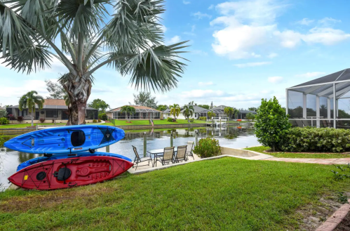 Two kayaks, one red and one blue, are parked by a calm canal surrounded by lush greenery and houses.