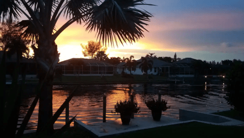 Sunset over a calm waterway, with palm trees and houses reflecting warm colors in the water.