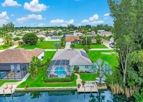 Aerial view of a residential area with houses, a pool, and lush greenery along a canal under a blue sky.