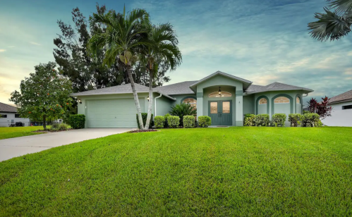 A charming green house with a manicured lawn and palm trees, set against a serene sky.