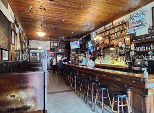 A cozy bar interior with wooden decor, a long counter, and shelves filled with bottles, featuring a few patrons.