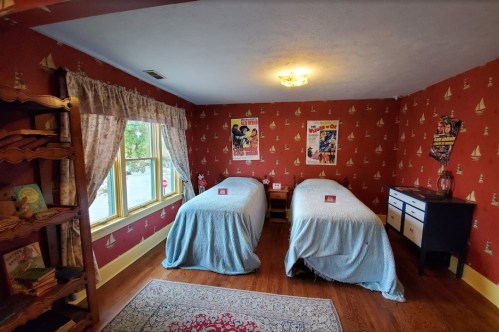 A cozy bedroom with two twin beds, a window, and vintage decor featuring sailboat wallpaper and classic posters.