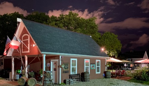 A charming red building at night, featuring a Texas flag and outdoor seating under a cloudy sky.