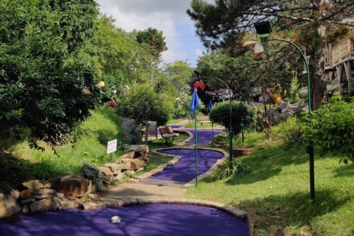 A winding mini-golf path with purple turf, surrounded by greenery and decorative plants under a cloudy sky.