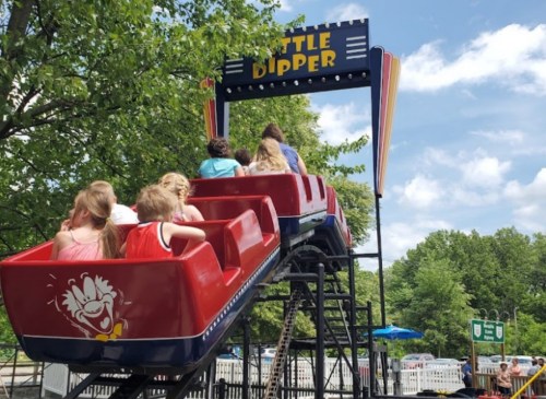 Children enjoy a ride on the Little Dipper roller coaster under a sunny sky, surrounded by trees.