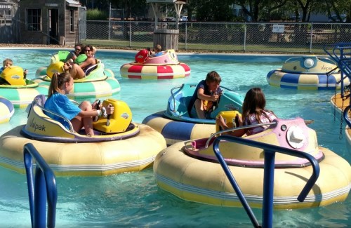 Children enjoying bumper boats in a sunny water park, surrounded by colorful boats and a clear blue pool.