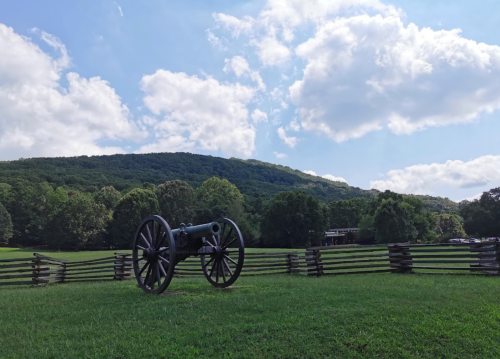 A historic cannon on a grassy field, surrounded by a wooden fence and hills under a partly cloudy sky.