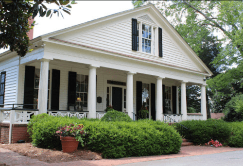 A charming white house with black shutters, a front porch, and lush greenery surrounding it.