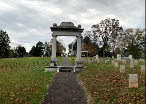 A stone archway leads to a cemetery with gravestones, surrounded by trees under a cloudy sky.