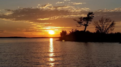 A serene sunset over a calm lake, with silhouettes of trees and clouds reflecting on the water's surface.