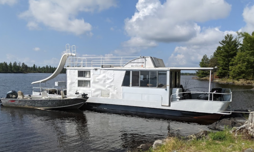 A white houseboat with a slide docked by a serene lake, surrounded by trees and blue skies.
