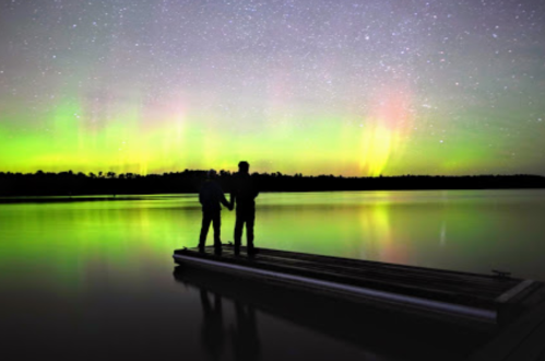 Two silhouetted figures stand hand in hand on a dock, gazing at vibrant auroras reflecting on a calm lake.