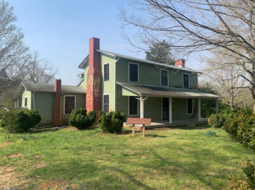 A green two-story house with a red chimney, surrounded by shrubs and trees, on a sunny day.