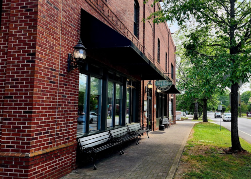 A brick building with large windows and benches outside, surrounded by trees and a sidewalk.