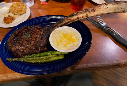 A large grilled ribeye steak with a long bone, served with asparagus and cheesy grits on a blue plate.