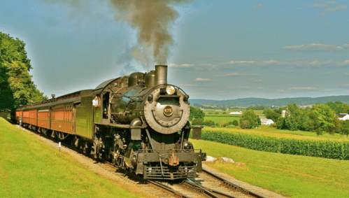 A vintage steam train chugs along a scenic countryside track, surrounded by green fields and blue skies.