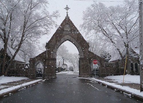 Snow-covered entrance to a stone archway with a cross, surrounded by trees, on a quiet, snowy street.