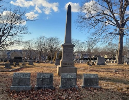 A cemetery scene featuring a tall obelisk monument surrounded by several gravestones under a clear blue sky.