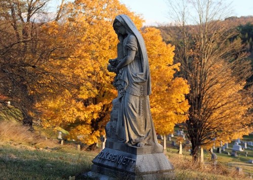A weathered statue of a woman holding flowers stands in a cemetery, framed by vibrant autumn trees.