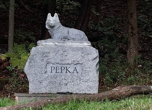A stone monument featuring a carved dog resting on a granite base with the inscription "РЕПКА." Surrounded by greenery.