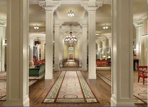 Elegant hotel lobby with tall columns, ornate chandeliers, and a patterned carpet leading to a reception area.